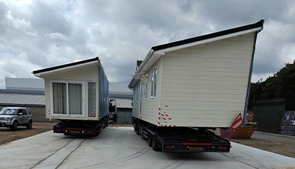 a truck transporting a mobile home on a road with trees and parked cars nearby while displaying house 3 features